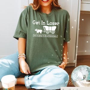 May include: A green t-shirt with a white graphic that says "Get in Loser, we're going to die of dysentery." The graphic is a pixelated image of a covered wagon being pulled by an ox.