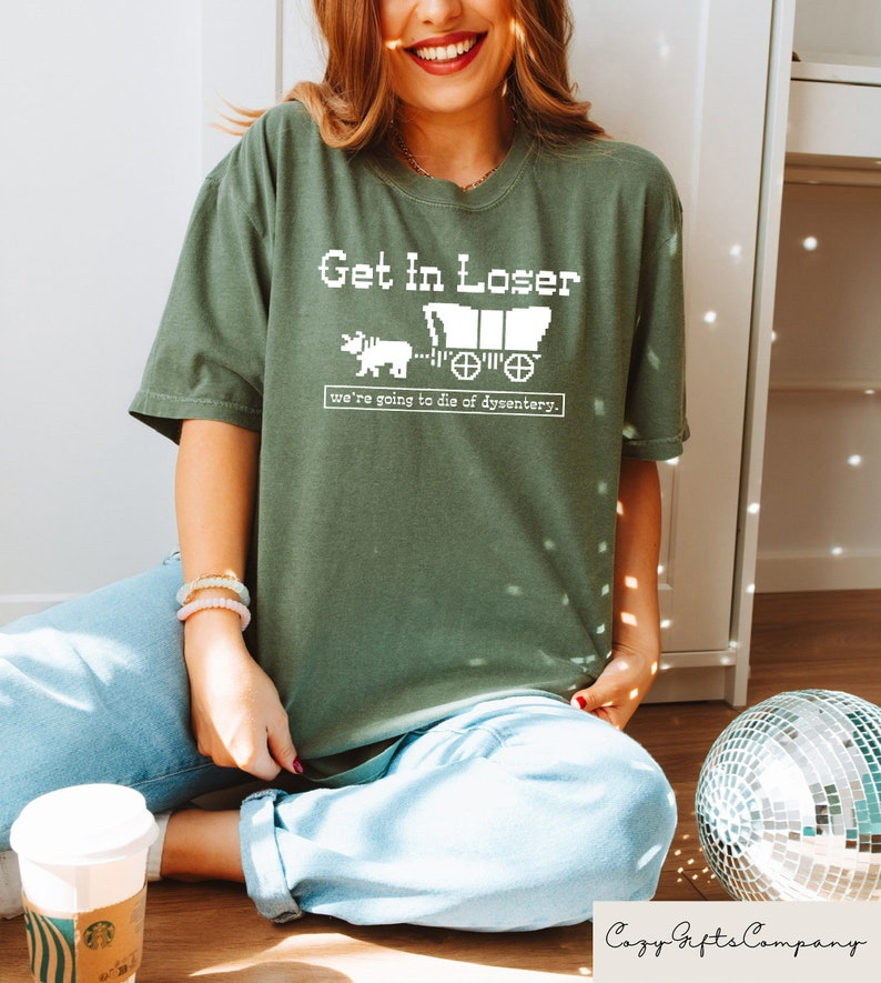 May include: A green t-shirt with a white graphic that says "Get in Loser, we're going to die of dysentery." The graphic is a pixelated image of a covered wagon being pulled by an ox.