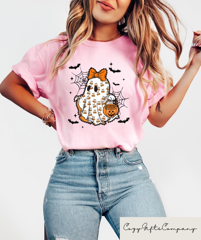 Puede incluir: Camiseta rosa claro con un dise&ntilde;o de fantasma de Halloween. El fantasma tiene un lazo naranja, lleva un cubo de calabaza y est&aacute; rodeado de murci&eacute;lagos y telara&ntilde;as. El texto "Cozy Gifts Company" est&aacute; en la parte inferior.