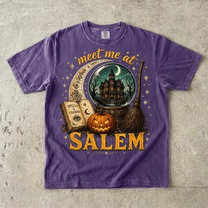 May include: A purple short-sleeve t-shirt featuring a graphic print with the text 'meet me at SALEM'. The design includes a crescent moon, a crystal ball showing a haunted house, an open book labeled 'SPELLS & CHARMS', a jack-o'-lantern, and a broom.