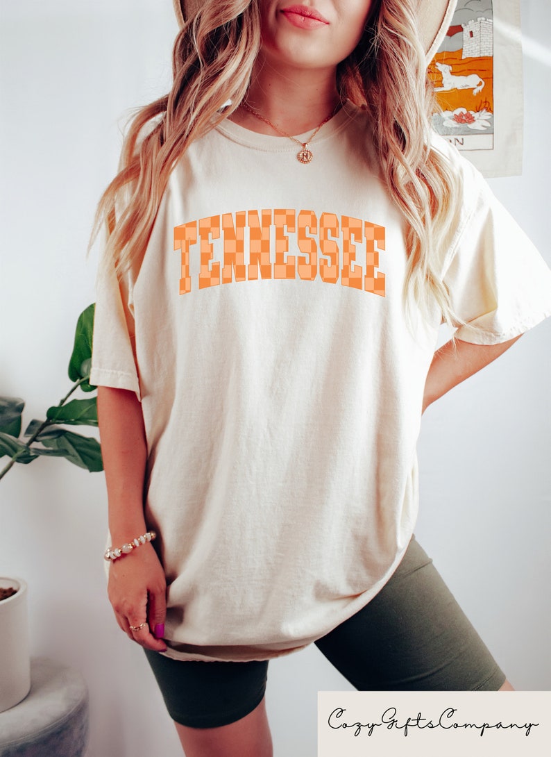 Pode incluir: Uma camiseta branca com um gr&aacute;fico de "Tennessee" xadrez laranja. A camiseta &eacute; de tamanho grande e tem um caimento solto.