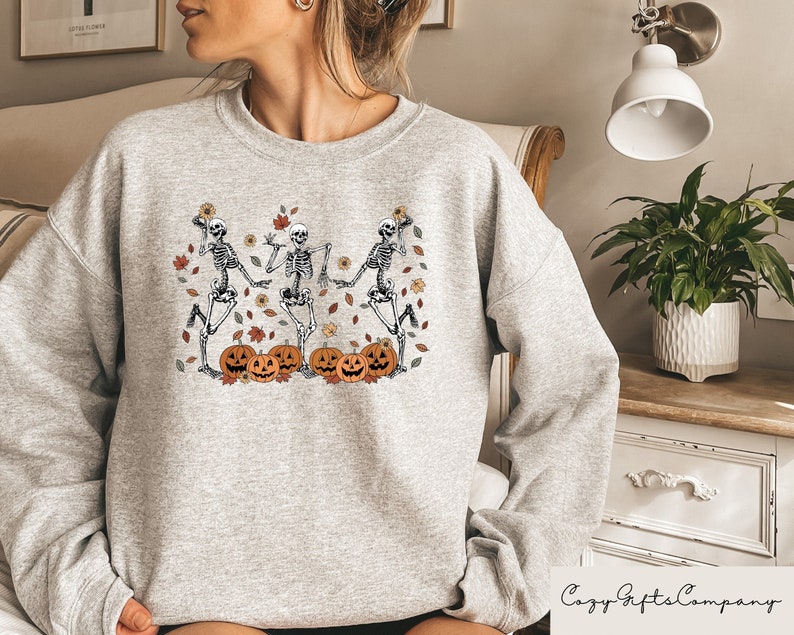 May include: Gray sweatshirt with a graphic of three skeletons dancing with pumpkins and fall leaves. The skeletons are wearing hats and holding pumpkins.