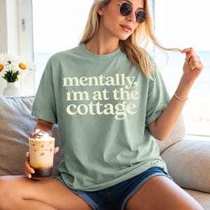 以下が含まれることがあります： 「mentally, I'm at the cottage」というクリーム色の文字が入ったセージグリーンのTシャツ。デニムのショートパンツと合わせています。ホイップクリームを添えたアイスコーヒーのグラスが手に持たれています。