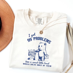 Puede incluir: Camiseta color crema con el texto "I got 99 PROBLEMS" y un oso de dibujos animados empujando un carrito de la compra. El texto "And a sweet little treat would solve most of them" está debajo del oso. Un collar de oro está sobre la camiseta.