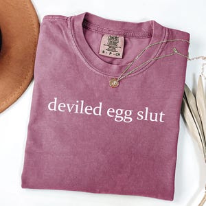 Puede incluir: Una camiseta rosa polvorienta con las palabras "deviled egg slut" impresas en blanco. Un collar dorado con un peque&ntilde;o colgante descansa sobre la camiseta. Un sombrero marr&oacute;n y una planta seca est&aacute;n en el fondo.