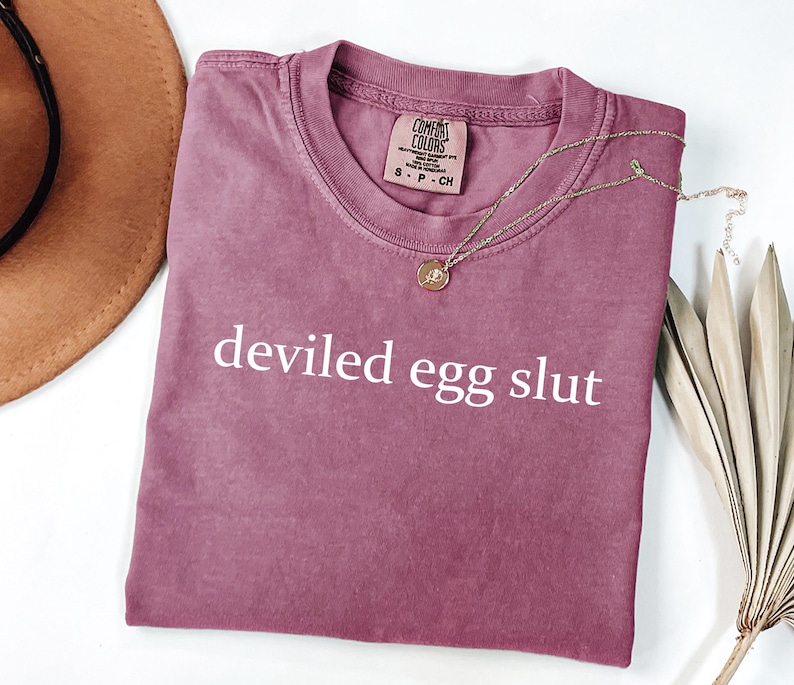 Puede incluir: Una camiseta rosa polvorienta con las palabras "deviled egg slut" impresas en blanco. Un collar dorado con un peque&ntilde;o colgante descansa sobre la camiseta. Un sombrero marr&oacute;n y una planta seca est&aacute;n en el fondo.