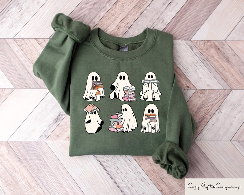 Puede incluir: Una sudadera verde con un gr&aacute;fico de seis fantasmas blancos sosteniendo pilas de libros. Los fantasmas est&aacute;n en varias poses, algunos est&aacute;n leyendo, algunos est&aacute;n sosteniendo los libros frente a sus caras y otros simplemente est&aacute;n sosteniendo los libros. El texto "Cozy Gifts Company" est&aacute; impreso en blanco en la esquina inferior derecha de la sudadera.