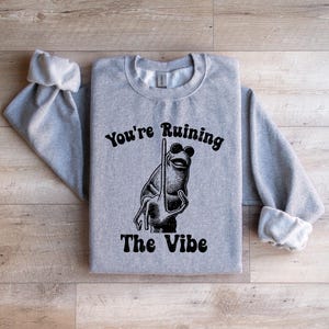 May include: A pale grey sweatshirt featuring the text "You're Ruining The Vibe" in black. A cartoon frog wearing sunglasses and holding a pole is in the centre. The sweatshirt has long sleeves with rolled cuffs.