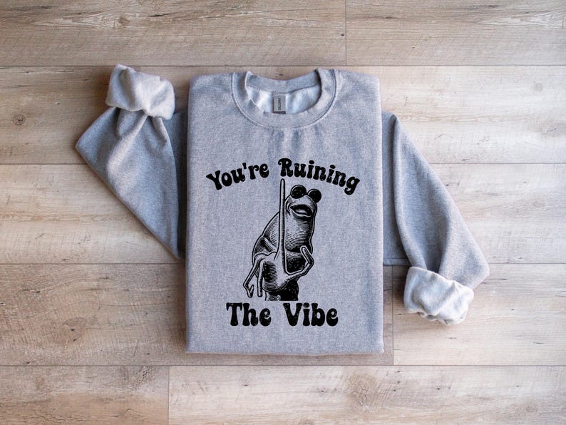 May include: A pale grey sweatshirt featuring the text "You're Ruining The Vibe" in black. A cartoon frog wearing sunglasses and holding a pole is in the centre. The sweatshirt has long sleeves with rolled cuffs.