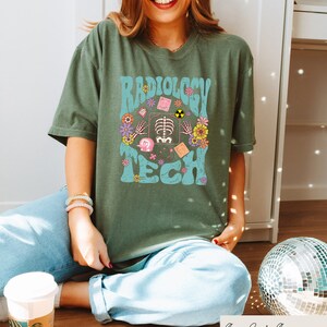 May include: A green t-shirt with a retro-style graphic design that says "Radiology Tech" in blue and pink lettering. The graphic includes a skeleton, flowers, and other medical symbols.