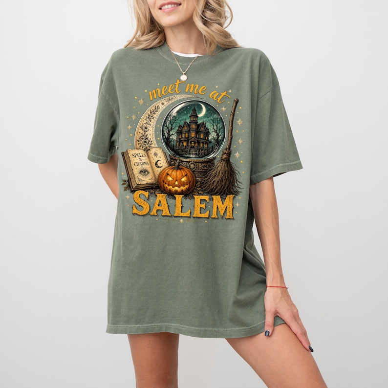May include: An olive green t-shirt featuring a graphic print of a crescent moon, a crystal ball with a haunted house, a spellbook labeled Spells & Charms, a jack-o'-lantern, and a broomstick, with the text meet me at SALEM printed in yellow.