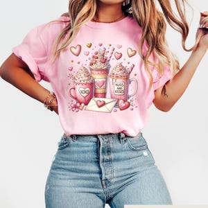 May include: Pink t-shirt featuring a Valentine's Day design with three coffee mugs, hearts, and an envelope. The mugs have whipped cream, hearts, and the phrases "XOXO" and "Hugs and Kisses". The shirt is paired with blue jeans.
