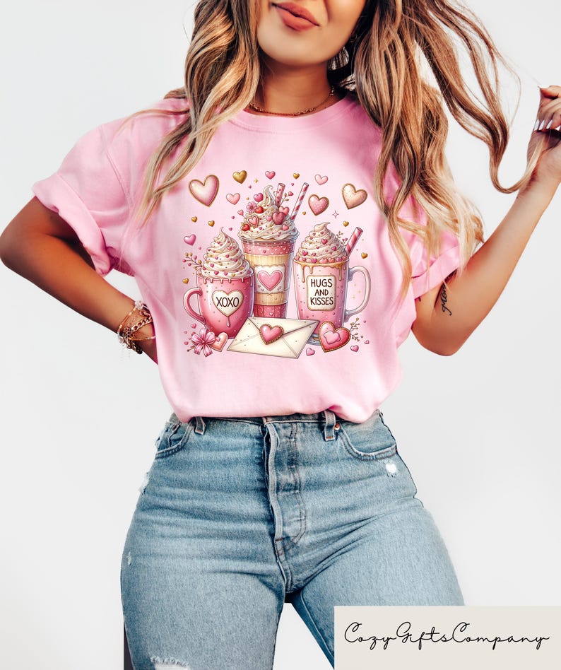 May include: Pink t-shirt featuring a Valentine's Day design with three coffee mugs, hearts, and an envelope. The mugs have whipped cream, hearts, and the phrases "XOXO" and "Hugs and Kisses". The shirt is paired with blue jeans.