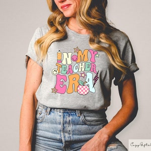 May include: A grey t-shirt featuring the text "IN MY TEACHER ERA" in a vibrant, retro style. The design incorporates a pencil, smiley face, stars, a lightning bolt, and a checkered apple. The shirt is a casual, short-sleeved design.
