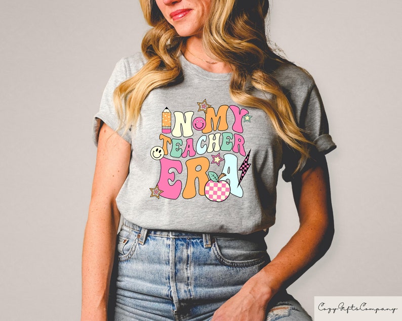 May include: A grey t-shirt featuring the text "IN MY TEACHER ERA" in a vibrant, retro style. The design incorporates a pencil, smiley face, stars, a lightning bolt, and a checkered apple. The shirt is a casual, short-sleeved design.