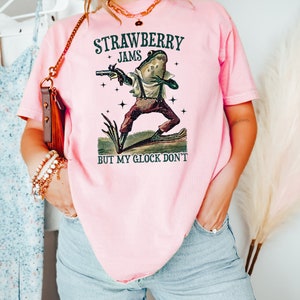 May include: Pink t-shirt with the text "STRAWBERRY JAMS BUT MY GLOCK DON'T" and a frog graphic holding a gun. The frog is wearing suspenders and pants. The shirt is a light pink color.
