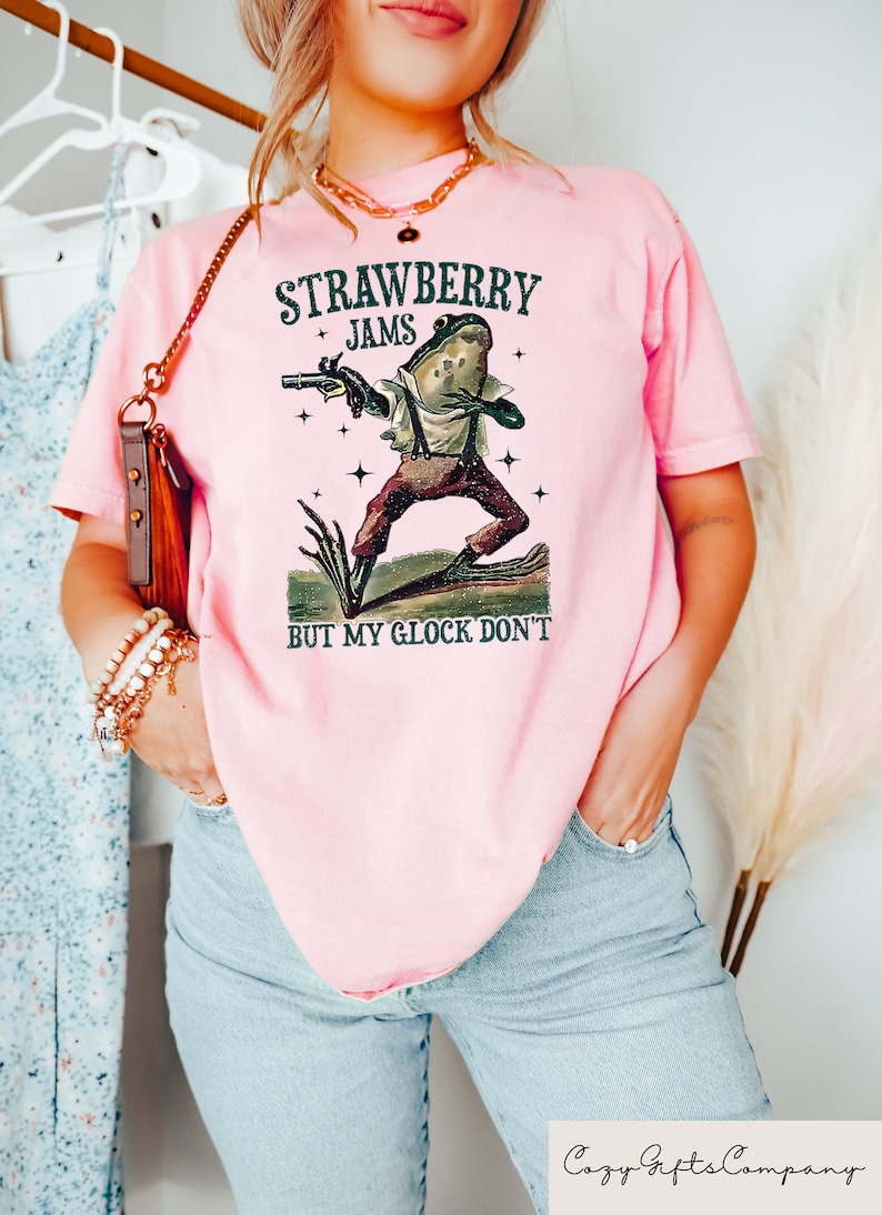 May include: Pink t-shirt with the text "STRAWBERRY JAMS BUT MY GLOCK DON'T" and a frog graphic holding a gun. The frog is wearing suspenders and pants. The shirt is a light pink color.