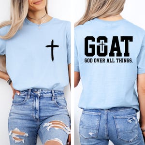 May include: Light blue t-shirt with a black cross on the front. The back of the shirt features the word "GOAT" with a cross in the "O", and the text "GOD OVER ALL THINGS."