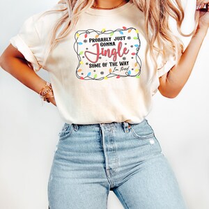 May include: A cream-coloured t-shirt featuring the text "Probably Just Gonna Jingle Some of the Way I'm Tired". The design includes colourful Christmas lights and snowflakes, ideal for the festive season.