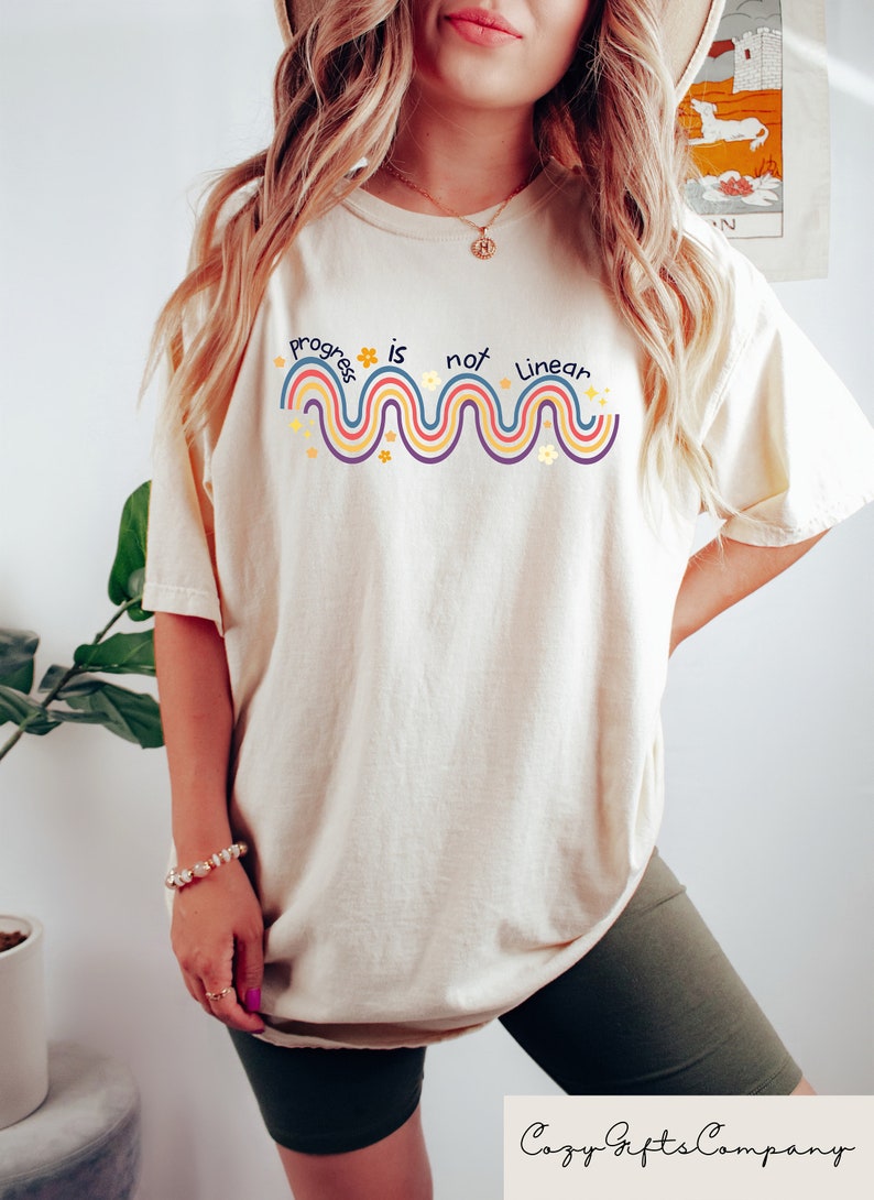 May include: A cream colored t-shirt with a graphic design that says "progress is not linear" with a wavy line and flowers in rainbow colors.