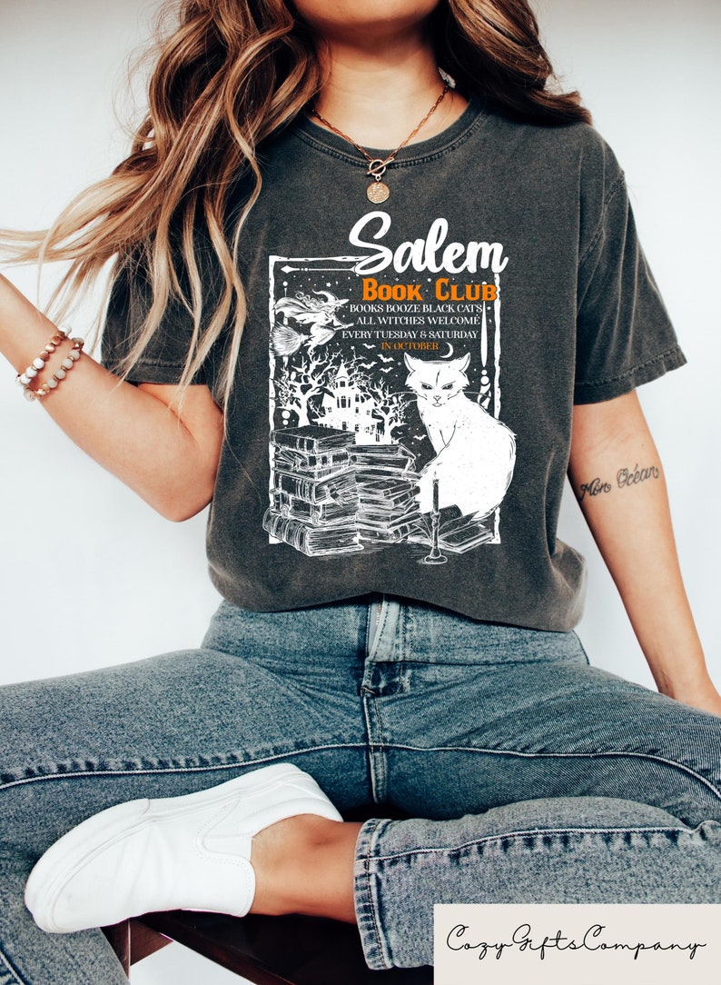 May include: Dark gray t-shirt with a white graphic featuring the text "Salem Book Club." The design includes a witch on a broom, a black cat, and a stack of books. The shirt also has the text "Books, Booze, Black Cats, All Witches Welcome."