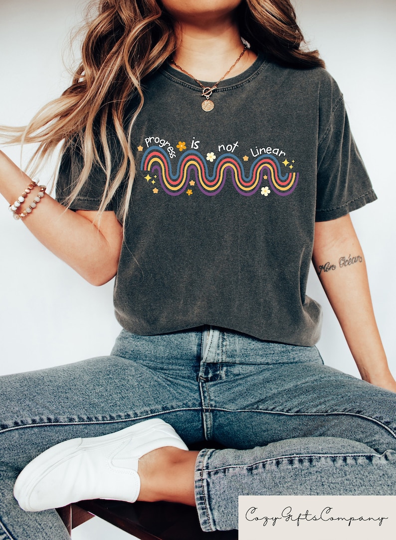 May include: A dark gray t-shirt with a colorful graphic that says "Progress is not linear" with a wavy line and stars and flowers.