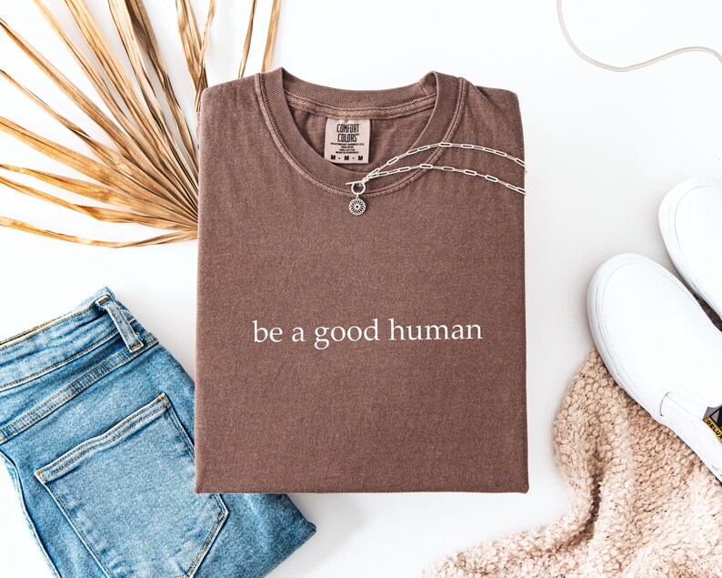 May include: A brown t-shirt with the text "be a good human" in white. The shirt is paired with blue jeans, white sneakers, and a silver necklace. A dried palm leaf adds a natural element to the composition.