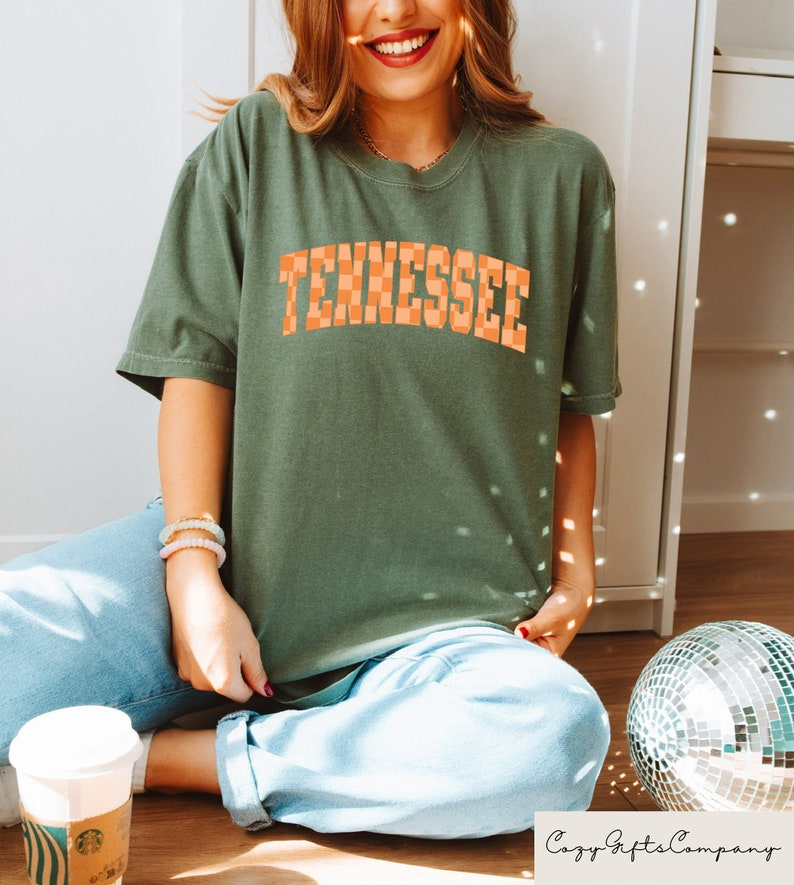 Pode incluir: Uma camiseta verde com a palavra "Tennessee" impressa em letras xadrez laranja.