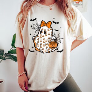 Puede incluir: Camiseta color crema con un dise&ntilde;o de fantasma de Halloween. El fantasma tiene un lazo naranja, detalles de calabaza y un cubo de linterna. Murci&eacute;lagos negros y telara&ntilde;as rodean al fantasma. El texto "Cozy Gifts Company" est&aacute; en la parte inferior.
