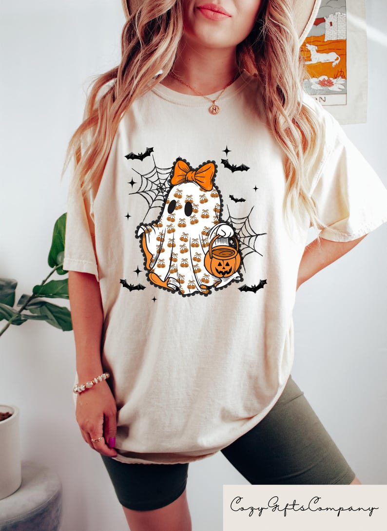 Puede incluir: Camiseta color crema con un dise&ntilde;o de fantasma de Halloween. El fantasma tiene un lazo naranja, detalles de calabaza y un cubo de linterna. Murci&eacute;lagos negros y telara&ntilde;as rodean al fantasma. El texto "Cozy Gifts Company" est&aacute; en la parte inferior.