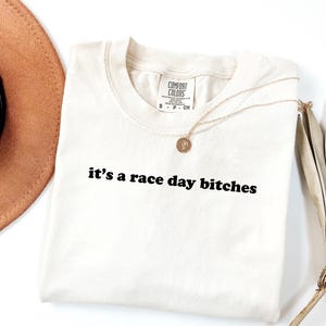 May include: Off-white t-shirt with the text "it's a race day bitches" in black. A gold necklace with a pendant is on the shirt. A brown hat and dried palm leaves are partially visible.
