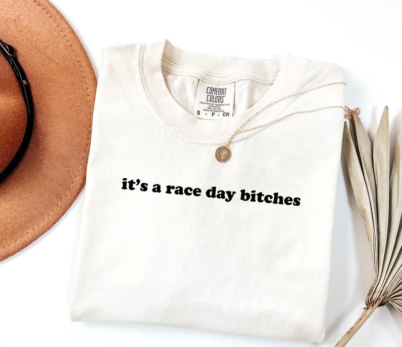 May include: Off-white t-shirt with the text "it's a race day bitches" in black. A gold necklace with a pendant is on the shirt. A brown hat and dried palm leaves are partially visible.