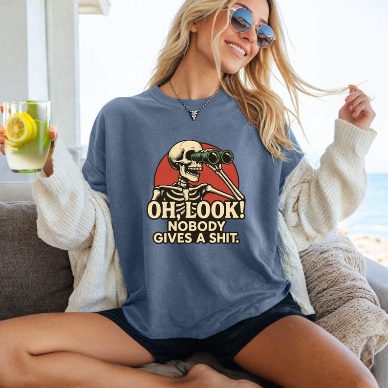 Puede incluir: Una camiseta azul con un gr&aacute;fico de un esqueleto mirando a trav&eacute;s de binoculares, sobre un c&iacute;rculo rojo. El texto dice "OH LOOK! NOBODY GIVES A SHIT." La camiseta la lleva una persona.