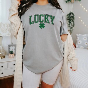K&ouml;nnte beinhalten: Graues T-Shirt mit dem Wort "LUCKY" in gr&uuml;nen Buchstaben und einem gr&uuml;nen Kleeblatt. Das Shirt wird mit wei&szlig;en gerippten Radlerhosen und einer cremefarbenen Strickjacke kombiniert.