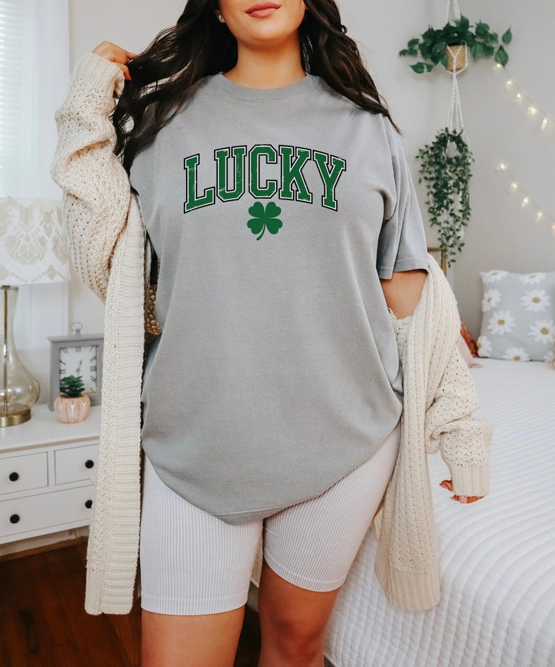 K&ouml;nnte beinhalten: Graues T-Shirt mit dem Wort "LUCKY" in gr&uuml;nen Buchstaben und einem gr&uuml;nen Kleeblatt. Das Shirt wird mit wei&szlig;en gerippten Radlerhosen und einer cremefarbenen Strickjacke kombiniert.