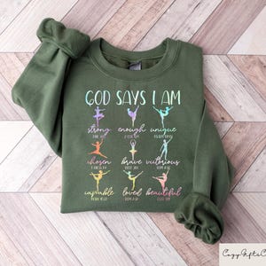 May include: An olive green sweatshirt with the text "GOD SAYS I AM" and the words "strong, enough, unique, chosen, brave, victorious, capable, loved, beautiful" in a colorful, stylized font. The design includes ballet dancer silhouettes. The Cozy Gifts Company logo is in the bottom right.