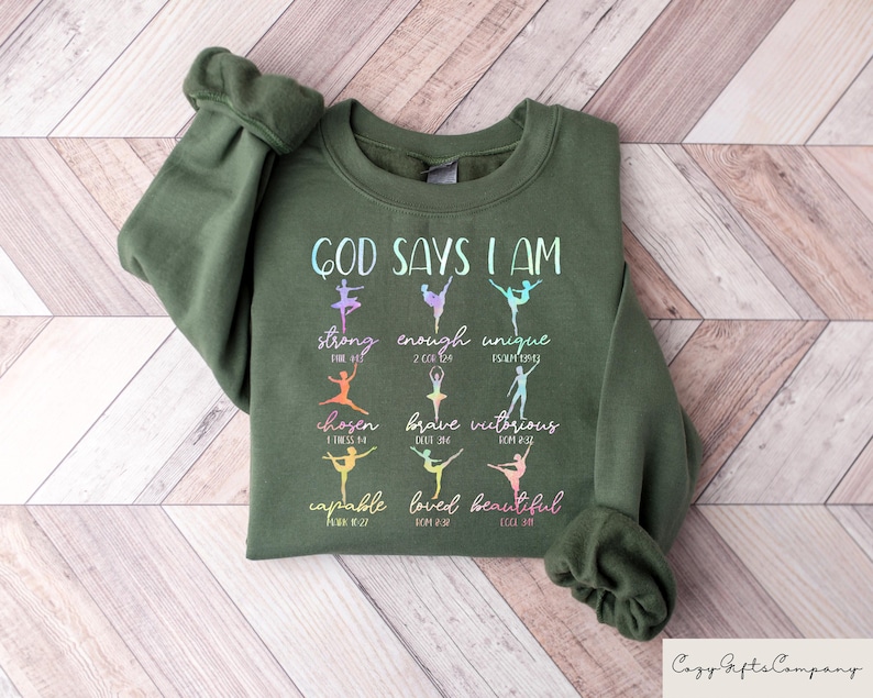 May include: An olive green sweatshirt with the text "GOD SAYS I AM" and the words "strong, enough, unique, chosen, brave, victorious, capable, loved, beautiful" in a colorful, stylized font. The design includes ballet dancer silhouettes. The Cozy Gifts Company logo is in the bottom right.