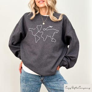 Puede incluir: Una sudadera gris oscuro con un contorno blanco de un mapa del mundo y un peque&ntilde;o avi&oacute;n volando por encima. El texto "Cozy Gifts Company" es visible en la esquina inferior derecha de la imagen.