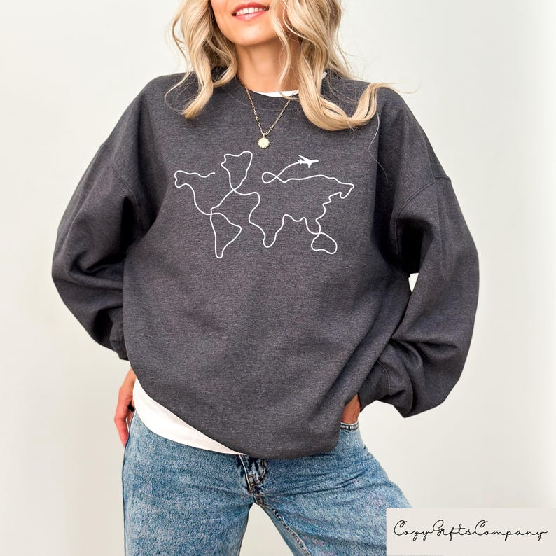 Puede incluir: Una sudadera gris oscuro con un contorno blanco de un mapa del mundo y un peque&ntilde;o avi&oacute;n volando por encima. El texto "Cozy Gifts Company" es visible en la esquina inferior derecha de la imagen.