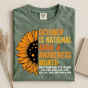 Könnte beinhalten: Ein salbeigrünes T-Shirt mit einer großen gelben Sonnenblume auf der linken Seite. Der Text lautet: OCTOBER IS NATIONAL ADHD AWARENESS MONTH BUT FOR ME IT'S ALSO JAN, FEB, MAR, APR, MAY, JUN, JUL, AUG, SEP, NOV & DEC, inklusive einer orangefarbenen Schleife.