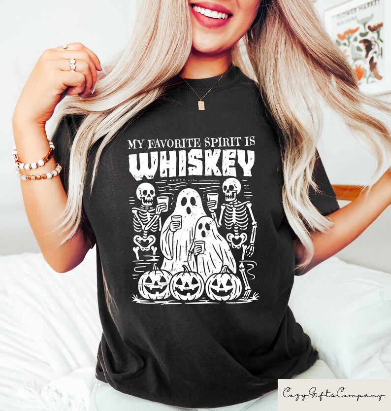 May include: Black t-shirt with the text "MY FAVORITE SPIRIT IS WHISKEY" in white. The graphic features skeletons, ghosts, and jack-o'-lanterns, perfect for Halloween. The shirt is a comfortable, casual wear.