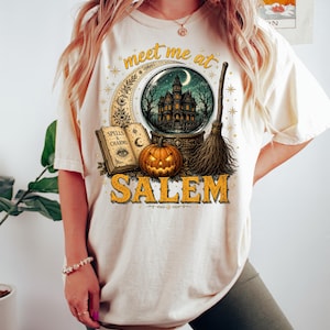 May include: A cream-colored t-shirt featuring a graphic print with the text "meet me at SALEM". The design includes a crescent moon, a crystal ball containing a haunted house, an open book labeled "SPELLS & CHARMS", a jack-o'-lantern, and a broom.