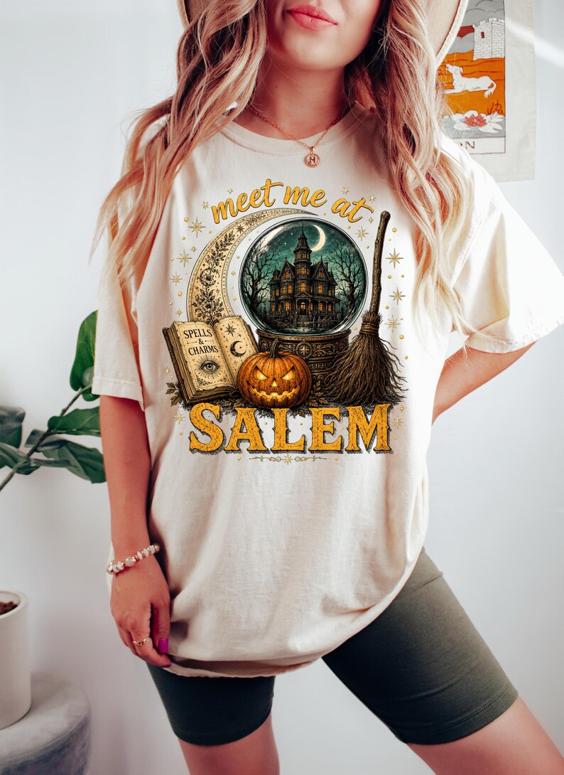 May include: A cream-colored t-shirt featuring a graphic print with the text "meet me at SALEM". The design includes a crescent moon, a crystal ball containing a haunted house, an open book labeled "SPELLS & CHARMS", a jack-o'-lantern, and a broom.
