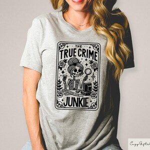 May include: A grey t-shirt featuring a black and white graphic of a skeleton holding a magnifying glass and a glass of wine. The graphic is in the shape of a playing card and reads "The True Crime Junkie".