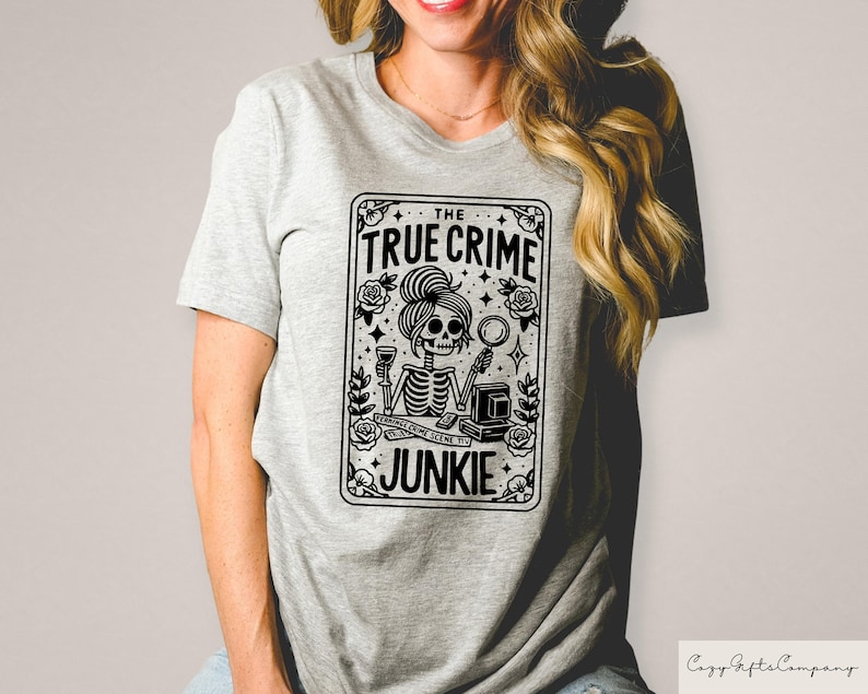 May include: A grey t-shirt featuring a black and white graphic of a skeleton holding a magnifying glass and a glass of wine. The graphic is in the shape of a playing card and reads "The True Crime Junkie".