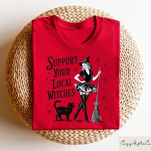 May include: Red t-shirt with a black and white illustration of a witch holding a broom. The text "Support Your Local Witches" is printed on the shirt.