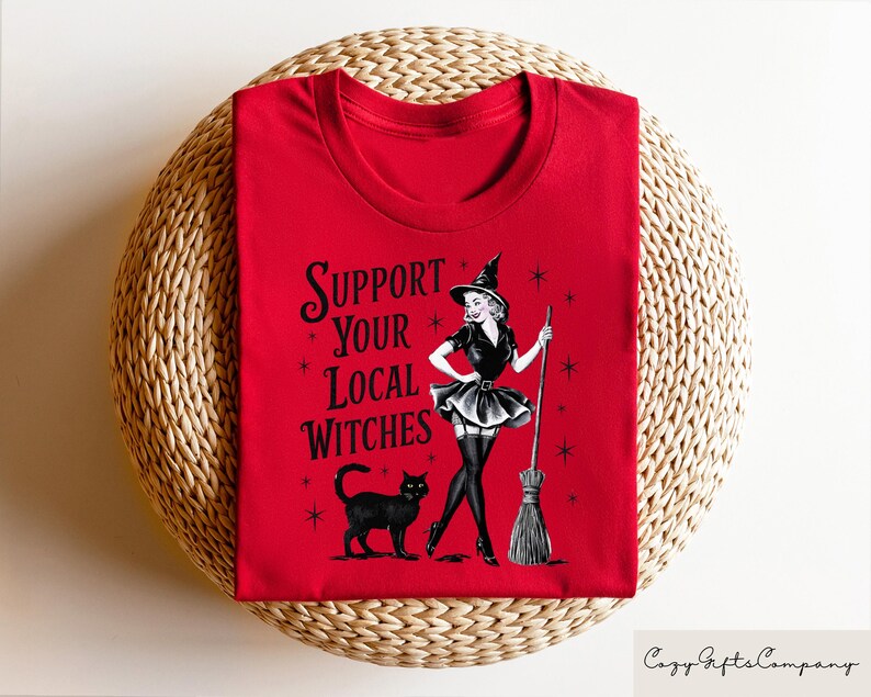 May include: Red t-shirt with a black and white illustration of a witch holding a broom. The text "Support Your Local Witches" is printed on the shirt.