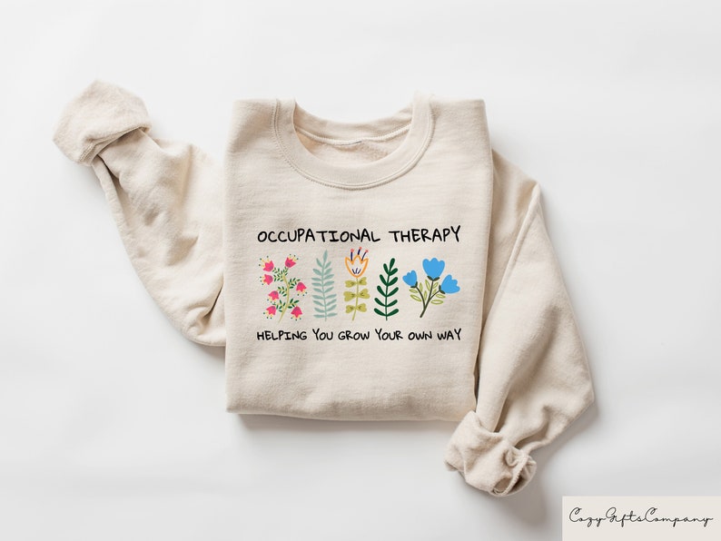 以下が含まれることがあります： 「Occupational Therapy」という文字と、「Helping you grow your own way」という文字が書かれたカラフルな花が並んだベージュのトレーナーです。