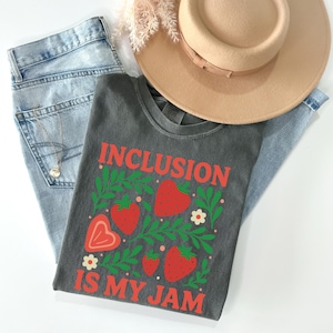 May include: A dark gray t-shirt with the words "INCLUSION IS MY JAM" in red, along with red strawberries and green leaves. The shirt is paired with light blue jeans and a tan hat.