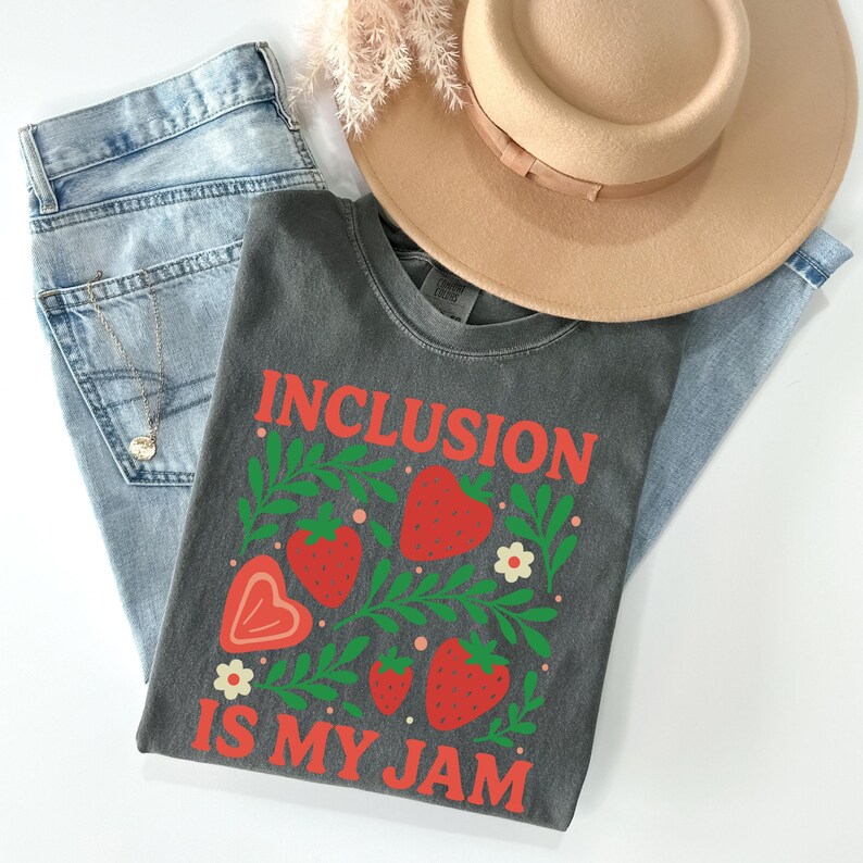 May include: A dark gray t-shirt with the words "INCLUSION IS MY JAM" in red, along with red strawberries and green leaves. The shirt is paired with light blue jeans and a tan hat.